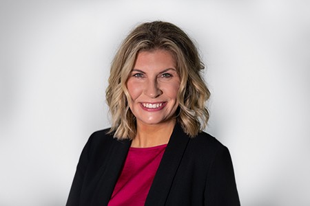 Bridgett Adams, a fair woman wearing a pink top with black blazer, short blond wavy hair, pink lipstick and big smile