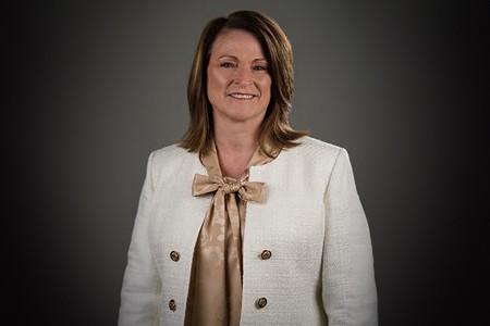 Pam Thompson, a white woman wearing light golden top, big bow and white blazer, short light brown hair and smile